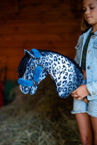 Lifestyle photo: a girl in a denim jacket holds the Hobby Horse Picasso with Black Halter by Hobby Horse LarDen against a red wooden barn wall - blue and white leopard-print plush head, black yarn mane - shown here with the fur-lined Teddy Petrol halter.