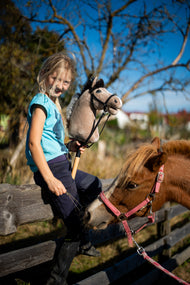 Blond dziewczynka w turkusowej bluzce siedzi na drewnianym ogrodzeniu padoku, trzymając Hobby Horse Piccolino z Black Halter od Hobby Horse LarDen, z jasnobeżową pluszową głową i ciemną włóczkową grzywą, z kasztanowatym kucykiem za nią, z czarnym ogłowiem Complete ze skóry ekologicznej