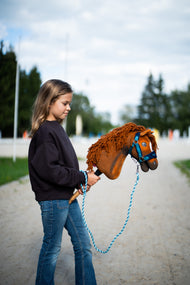 Dziewczyna w ciemnej bluzie prowadzi Hobby Horse Pippa z kantarem Black Halter od Hobby Horse LarDen piaszczystą ścieżką obok placu – kasztanowata pluszowa głowa, ruda grzywa z włóczki – pokazany z podszytym futerkiem kantarem LarDen Blue i Uwiązem Bloom Blue