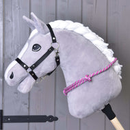 Cordeo w różowe paski Hobby Horse LarDen na szarej pluszowej głowie Hobby Horse z białą grzywą, czarnym ogłowiem i różowo-białym cordeo na drewnianym kiju, idealne do ćwiczenia jazdy bez ogłowia, na tle szarej drewnianej ściany.