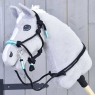 Hobby Horse LarDen Zestaw sznurkowy Turquoise pokazany na szarym pluszowym Hobby Horse z białą grzywą, czarnym ogłowiem i turkusowymi akcentami, osadzonym na drewnianym kiju przed szarą drewnianą ścianą.