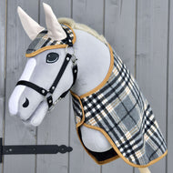 Hobby Horse LarDen Zestaw England Szary przedstawia szarego pluszowego Hobby Horse z czarną uzdą i wodzami oraz derką w kratę i dopasowanym nausznikiem, tworząc urokliwą dekorację na tle drewna.