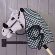 Zestaw Harmony Green od Hobby Horse LarDen — pluszowy Hobby Horse z czarnym kantarem, zielono-fioletową derką stajenną w pepitkę i zwiniętym czarnym sznurkiem, zawieszony na drewnianym uchwycie ściennym na tle szarej drewnianej ściany.
