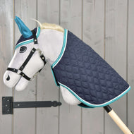 Hobby Horse LarDen Zestaw Supreme Turkusowy na szarym pluszowym koniku na kiju z niebieskimi uszami, czarną uzdą i pikowaną granatową derką z turkusowym wykończeniem, na tle jasnej drewnianej ściany.