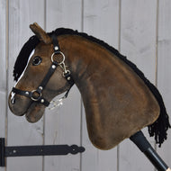 Hobby Horse Sundance z czarnym kantarem od Hobby Horse LarDen o pluszowym wykończeniu, z czarną grzywą z włóczki, uzdą i wodzami — oddający żywy wygląd ogiera rasy pełnej krwi angielskiej — osadzony na kiju, pokazany na tle ściany z jasnych drewnianych desek.