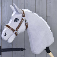 Hobby Horse LarDen Kantar ze sztucznym futrem - skóra ekologiczna Walnut to szary pluszowy Hobby Horse z białą grzywą, brązową uzdą ze skóry ekologicznej i dużymi czarnymi oczami na drewnianym kiju, pokazany oparty o szarą drewnianą ścianę.