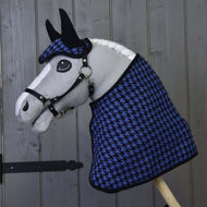 A Hobby Horse LarDen hobby horse with a white mane and black bridle wears the "Set festive checkered blue" (houndstooth-patterned ear bonnet and blanket) and poses before a gray wooden wall.