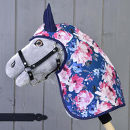 Hobby Horse LarDen „Płaszcz przeciwdeszczowy Peonia” to szary Hobby Horse z czarnym ogłowiem, ubrany w niebieski płaszcz przeciwdeszczowy ozdobiony różowymi i białymi kwiatami, osadzony na drewnianym kiju na tle szarej drewnianej ściany.