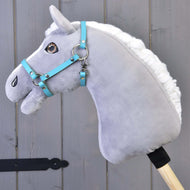 Szary pluszowy hobby horse z białą grzywą i niebieską uzdą ma założony Kantar - czarna skóra ekologiczna (rozmiar S) od Hobby Horse LarDen, osadzony na drewnianym kiju przed szarymi drewnianymi panelami.