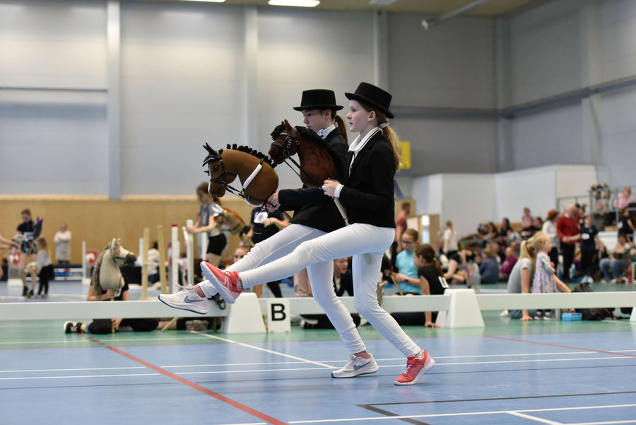 Dwoje dzieci w strojach zgodnych z formalnym dress code'em Hobby Horsing rywalizujących w hali z Hobby Horses