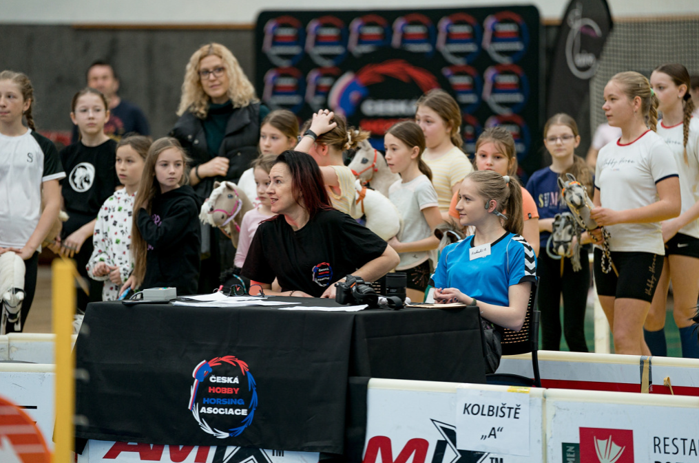 A busy indoor hobby horsing event scene with two organizers seated at a black table bearing the Česká Hobby Horsing Asociace logo, while girls in sportswear stand around them holding their hobby horses. White arena barriers, the sign Kolbiště „A“ and