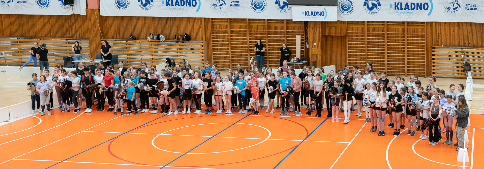 A large group of young hobby horsing competitors line up across an orange indoor sports court, wearing sporty casual clothes and holding many different hobby horses, with wooden wall bars, bleachers and Kladno banners in the background. The wide group