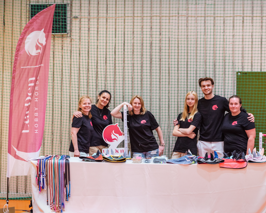 Six LarDen team members in matching black logo T-shirts pose behind a table of colorful reins, ribbons, and hobby-horsing accessories in an indoor sports hall, with a pink LarDen Hobby Horses banner and netting behind them. The friendly group stands