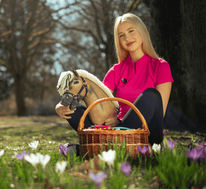Młoda kobieta w jaskraworóżowej koszulce siedzi na trawie na zewnątrz, trzymając zabawkowego Hobby Horse i wiklinowy koszyk wypełniony kolorowymi jajkami, otoczona kwitnącymi białymi i fioletowymi kwiatami.