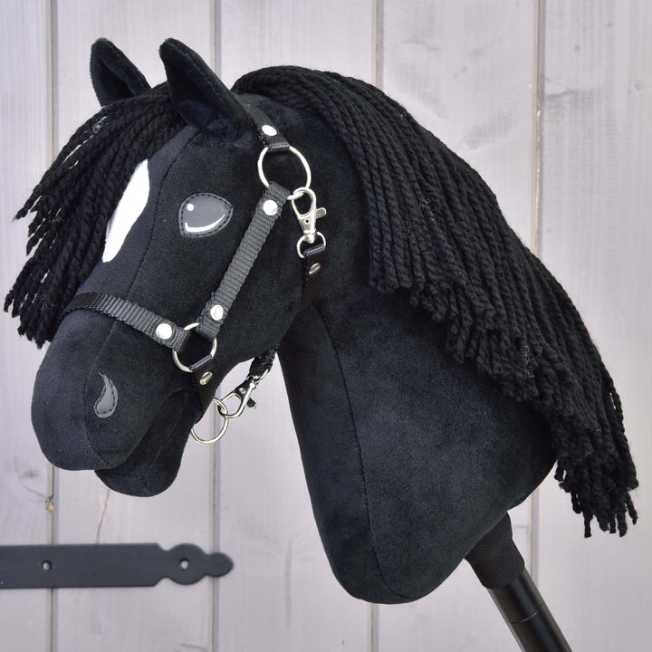 Czarny pluszowy Hobby Horse z długą włóczkową grzywą, białymi znaczeniami na pysku i czarną uzdą, osadzony na kiju, na tle jasnego drewnianego tła.