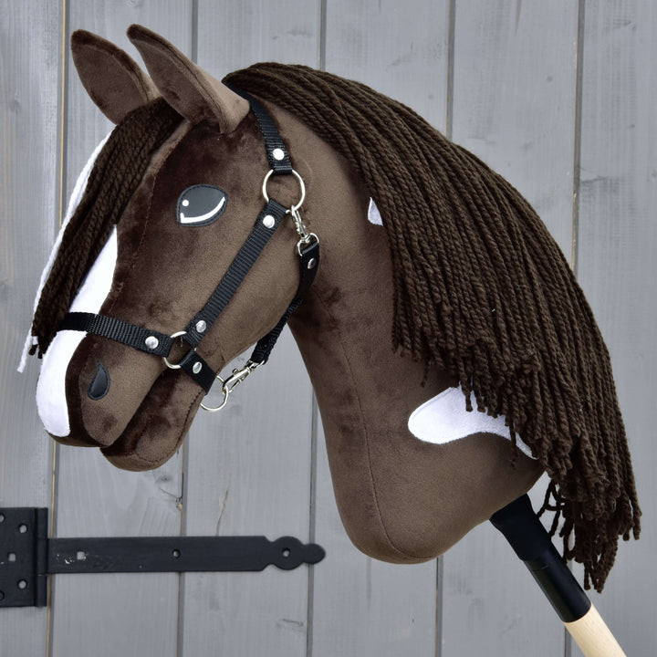 Realistyczny brązowo-biały Hobby Horse z czarną uzdą i długą włóczkową grzywą, osadzony na drewnianym kiju, ustawiony na tle szarego drewnianego płotu.