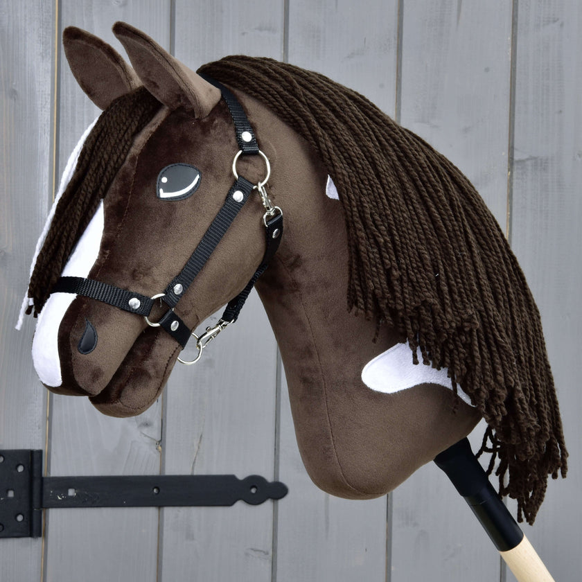 Realistyczny brązowo-biały Hobby Horse z czarną uzdą i długą włóczkową grzywą, osadzony na drewnianym kiju, ustawiony na tle szarego drewnianego płotu.