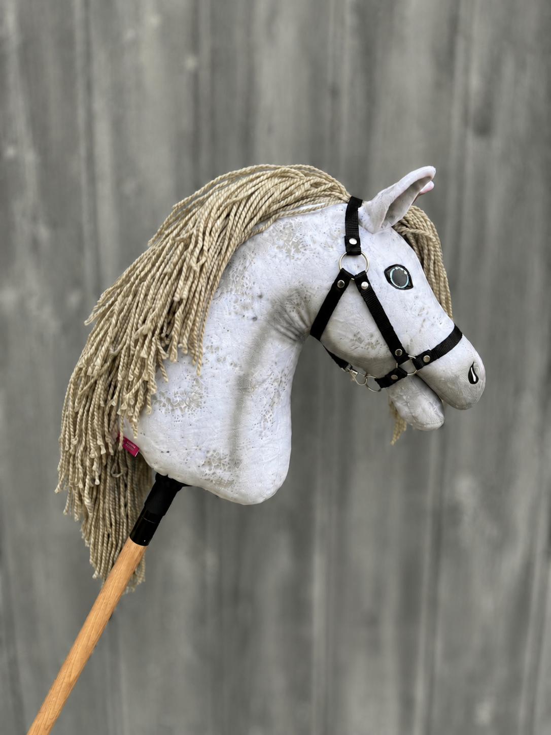 The Hobby Horse Victory with Black Halter (size M) by Hobby Horse LarDen features a white fabric head, yarn mane, black halter, and wooden stick—handmade and photographed against a gray wooden background.