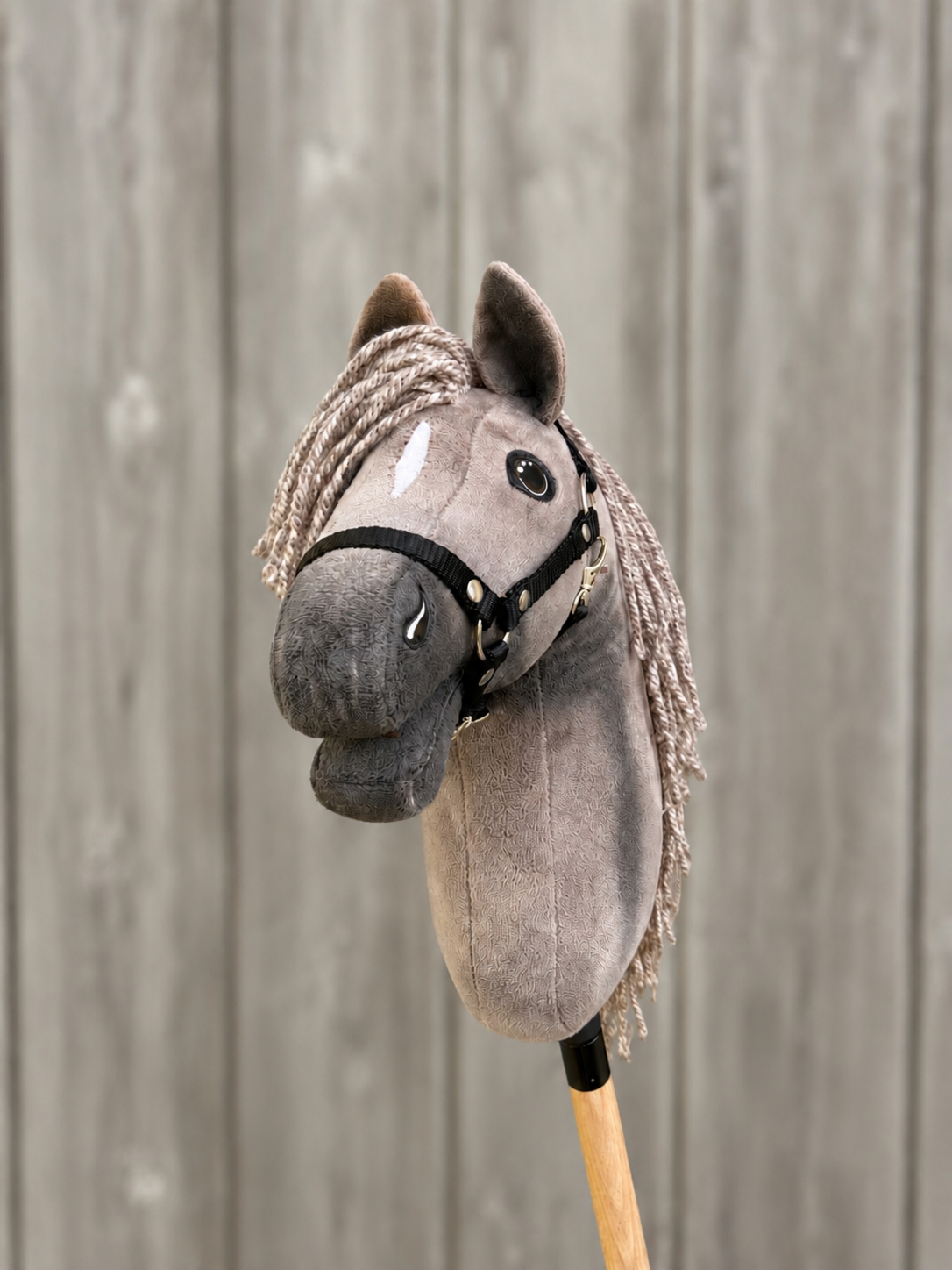 The Hobby Horse LarDen Hobby Horse Swing with Black Halter, size S, features a gray plush design on a wooden stick, set against a rustic wooden plank background.
