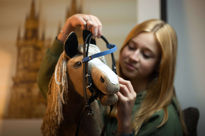 Młoda blond dziewczynka zakłada uzdę swojemu Hobby Horse, pluszowej zabawce z beżową grzywą. Jest skupiona i lekko się uśmiecha, ciesząc się sztuką Hobby Horsing w delikatnie rozmytym wnętrzu.