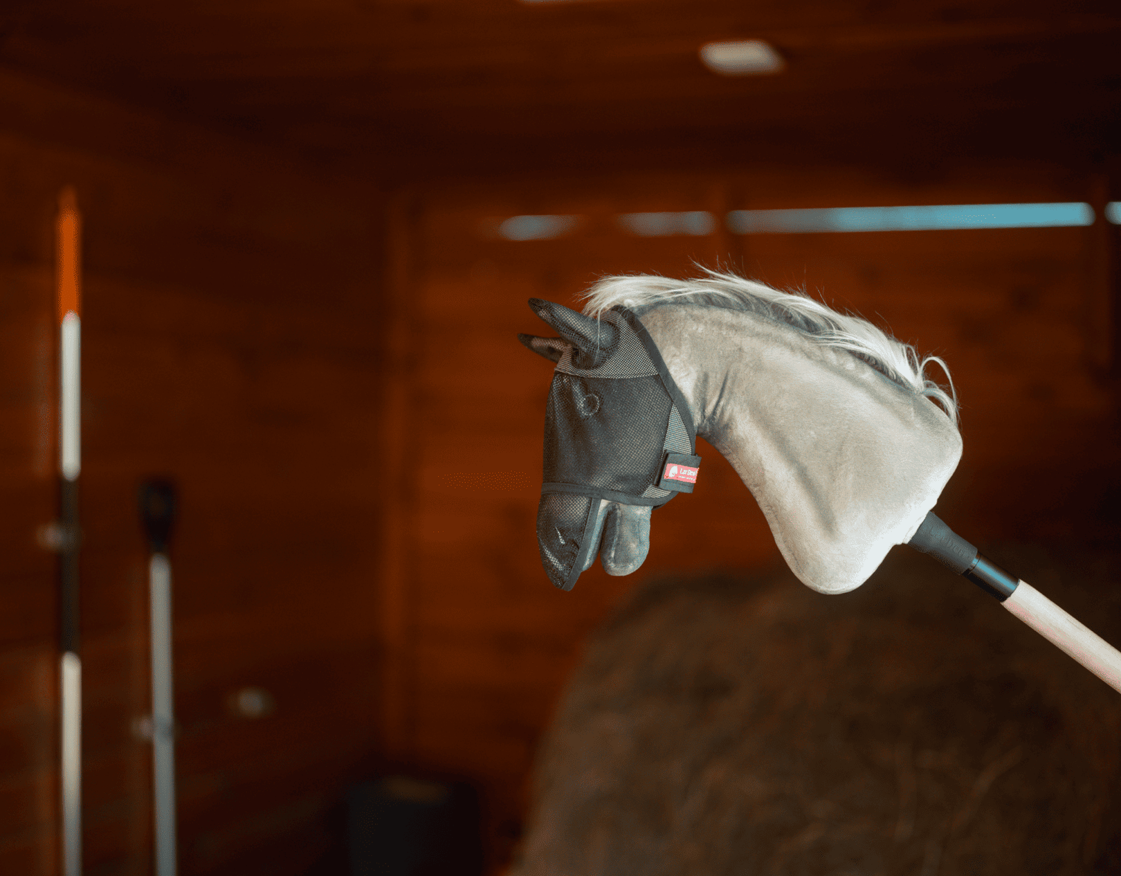 Hobby Horse z szarą pluszową głową i czarną uzdą jest osadzony na kiju wewnątrz drewnianej stodoły, w otoczeniu narzędzi gospodarskich i siana — idealny do pobudzania wyobraźni podczas przygód z Hobby Horsing.