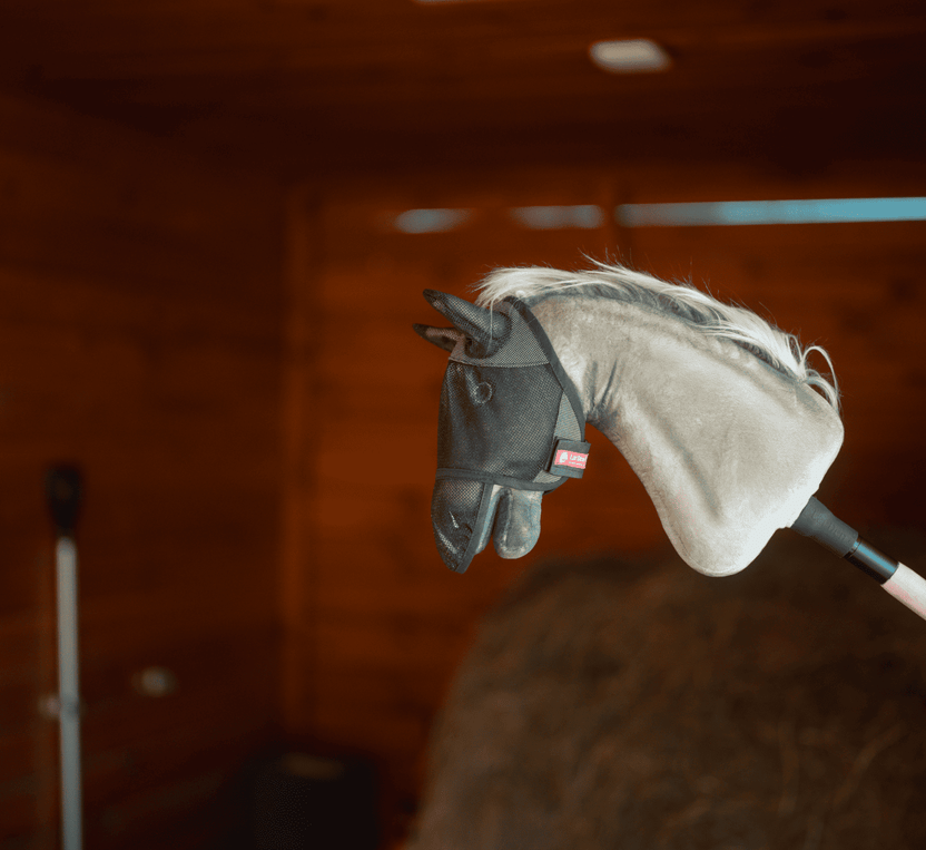 Pluszowa głowa Hobby Horse na kiju, w czarnej siatkowej masce przeciw owadom, pokazana we wnętrzu z drewnianymi ścianami i stosem siana — idealna do kreatywnej zabawy w Hobby Horsing.