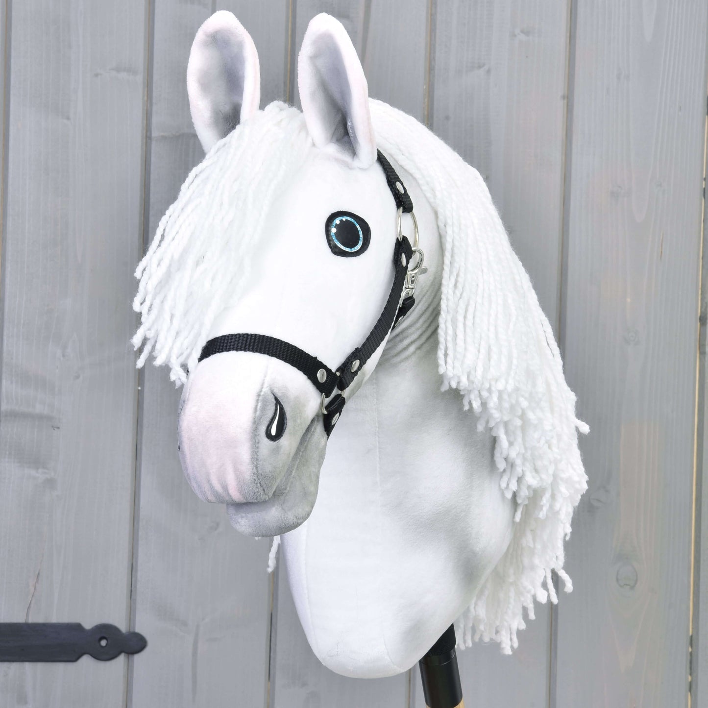 Hobby Horse Elektra z czarnym kantarem od Hobby Horse LarDen, ręcznie malowana biała klacz z długą grzywą z włóczki, niebieskimi oczami i czarnym kantarem, osadzona na kiju i pokazana na tle jasnoszarej drewnianej ściany.