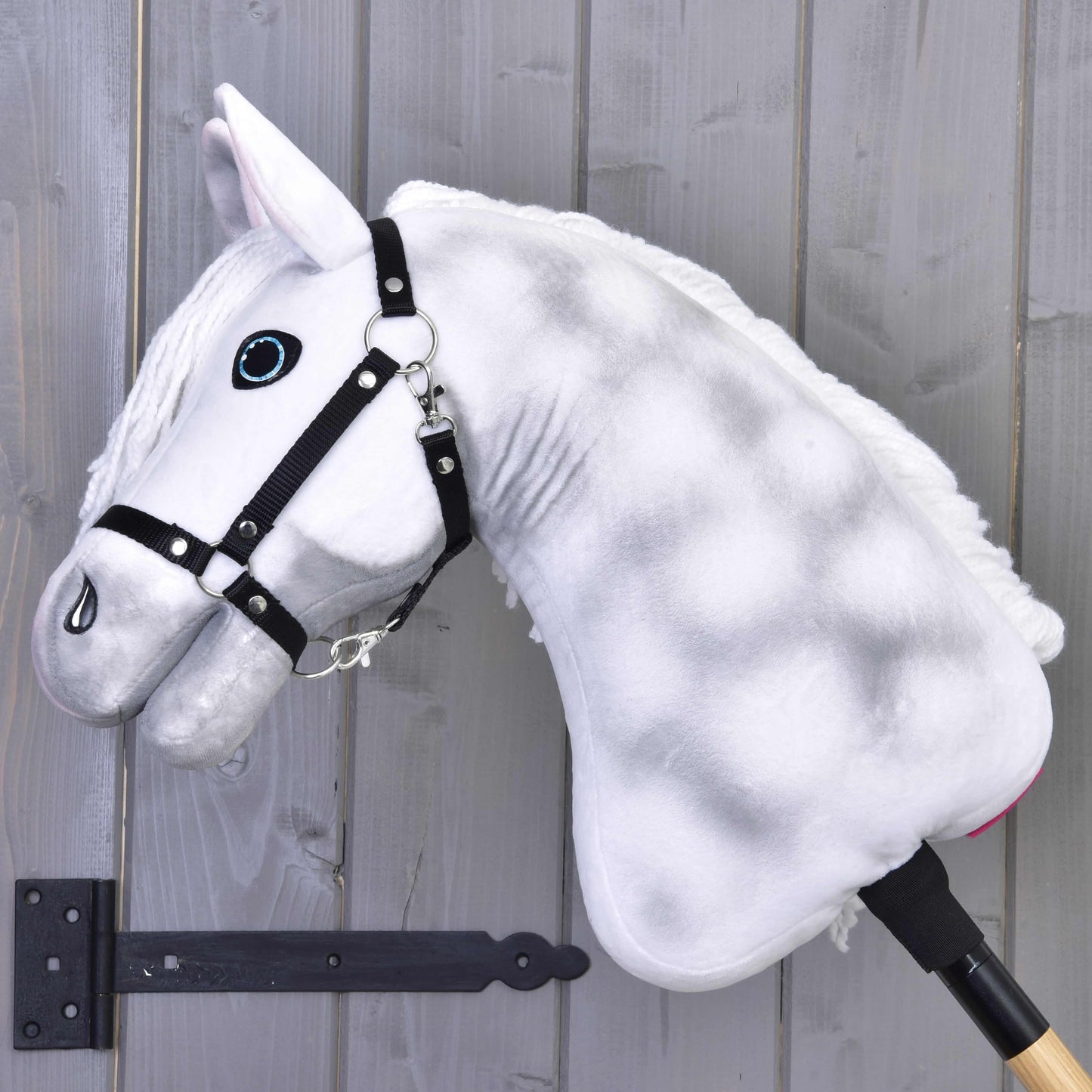 Hobby Horse Elektra z czarnym kantarem od Hobby Horse LarDen, z pluszowym białym wykończeniem, niebieskimi oczami i czarnym kantarem, osadzony na kiju, pokazany na tle szarej drewnianej ściany.