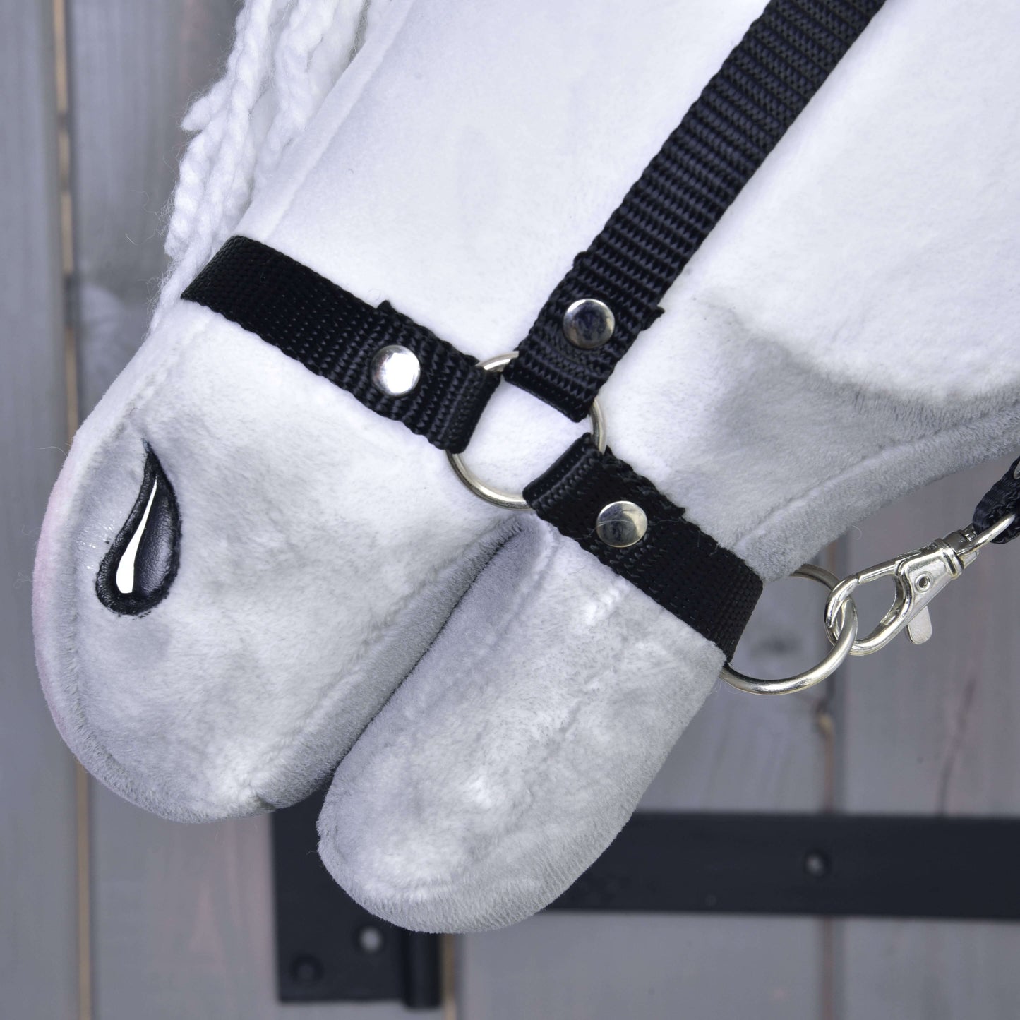 Zbliżenie na Hobby Horse Elektra z czarnym kantarem od Hobby Horse LarDen, pokazujące pluszowy biały pysk, czarny materiałowy kantar ze srebrnymi kółkami i klipsem oraz czarne szyte nozdrze, na tle drewna.