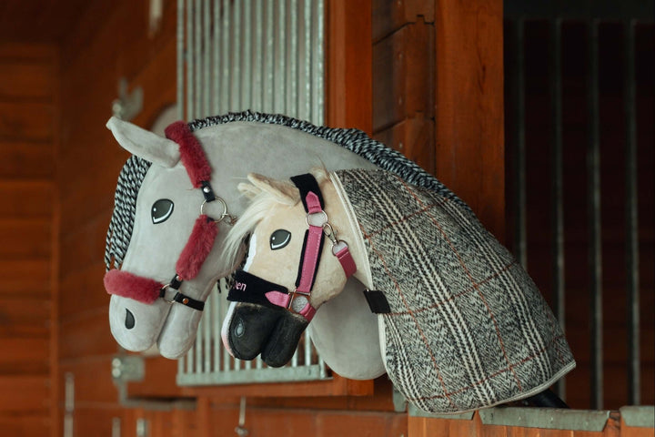 Dwie pluszowe zabawki hobby horse z uzdami i derkami spoczywają na drewnianej przegrodzie stajennej, przypominając prawdziwe konie w boksie. Idealne dla fanów hobby horsing, biały i szary koń mają dopracowane detale i rząd.