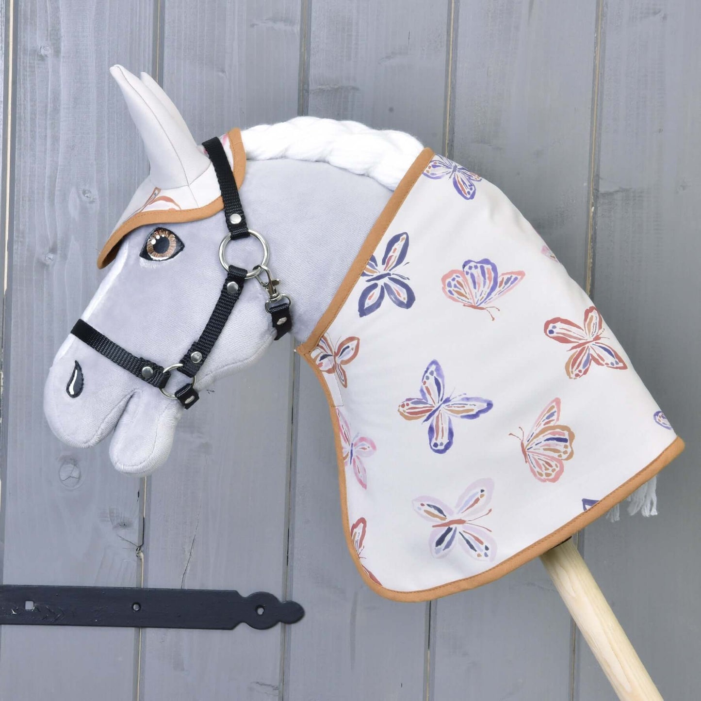 A grey hobby horse with a white mane wears a bridle and the Hobby Horse LarDen "Blanket Butterfly" softshell, featuring multicolored butterfly prints. Grey wooden paneling forms the background.
