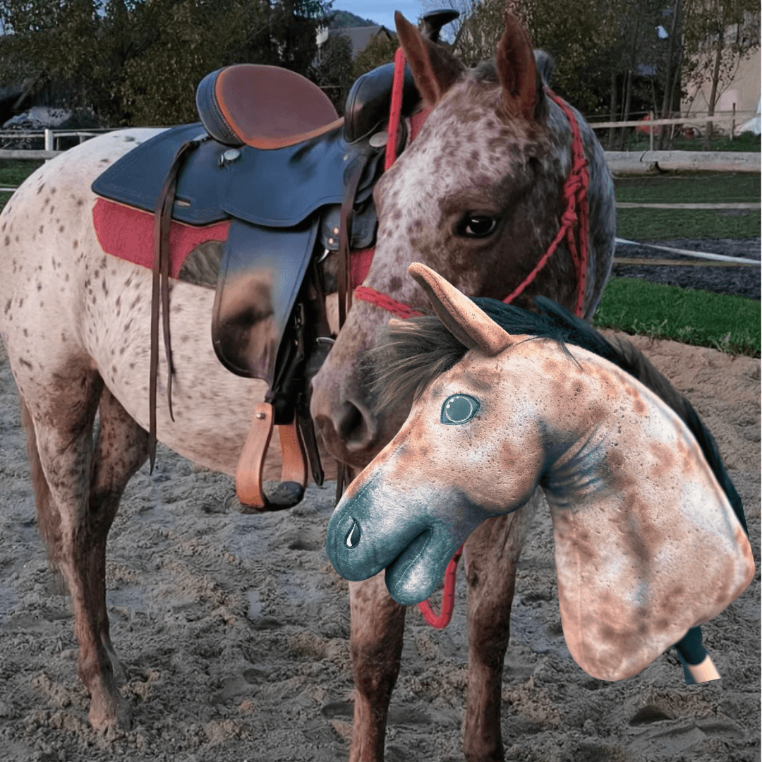A real brown and white spotted horse with a saddle stands on sand as someone holds a Hobby Horse LarDen "Hobby Horse based on your photo" near its face. Trees and fencing are visible in the background.