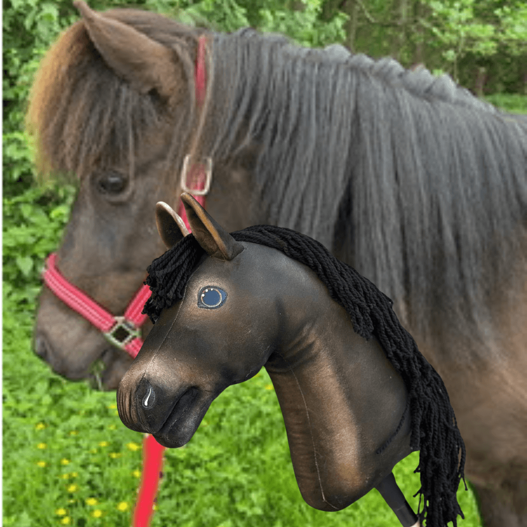 A real brown horse with a red halter stands outdoors beside a personalized "Hobby Horse based on your photo" by Hobby Horse LarDen—a brown stick horse with black yarn mane—set against greenery and trees.