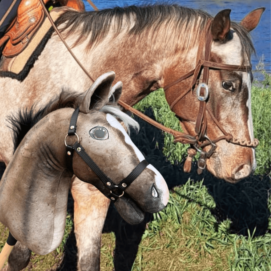 A person holds a Hobby Horse LarDen "Hobby Horse based on your photo" puppet, modeled after a real horse wearing a bridle, in front of the matching horse standing on grass near water.