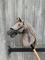 The Hobby Horse LarDen Hobby Horse Swing with Black Halter (size S), featuring a gray mane and expressive eyes, is mounted on a stick and rests against a weathered wooden door with visible grain and iron hardware.
