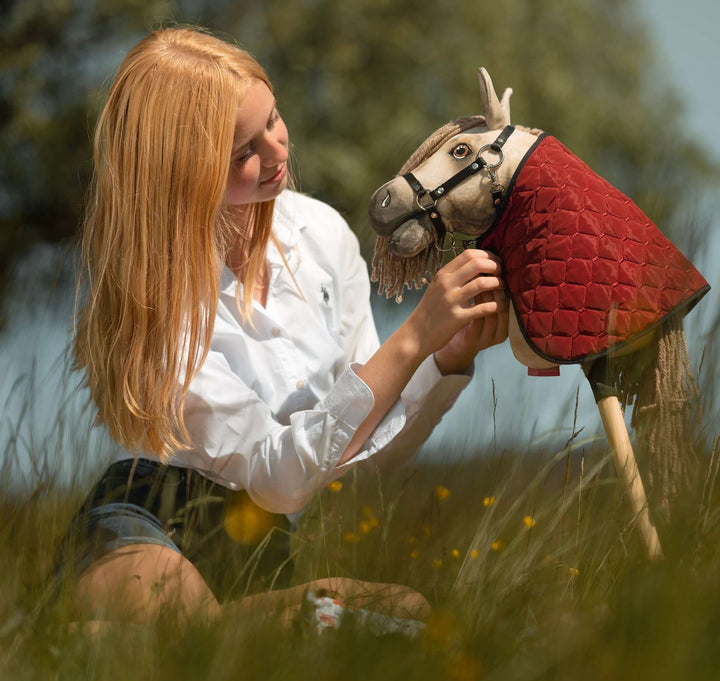 Młoda osoba z długimi blond włosami i w białej koszuli siedzi w wysokiej trawie, delikatnie poprawiając uzdę Hobby Horse z czerwoną pikowaną narzutą, ukazując pomysłowego ducha Hobby Horsing na tle rozmytej zieleni.