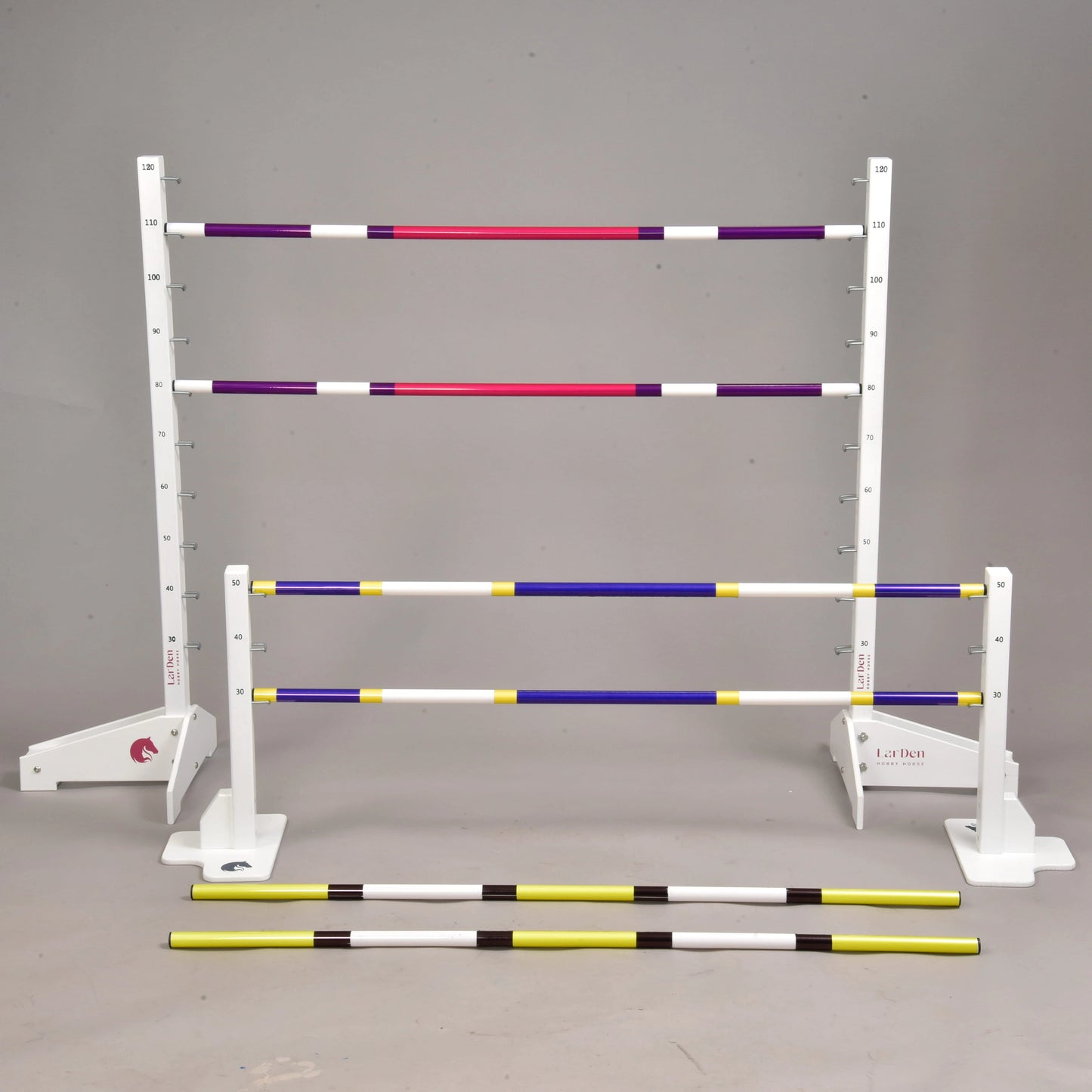 The Hobby Horse LarDen Jump set for Hobby Horsing is arranged in a studio, with adjustable jumps. Some striped purple, yellow, and white poles are set at various heights on stands, while others lie on the ground.