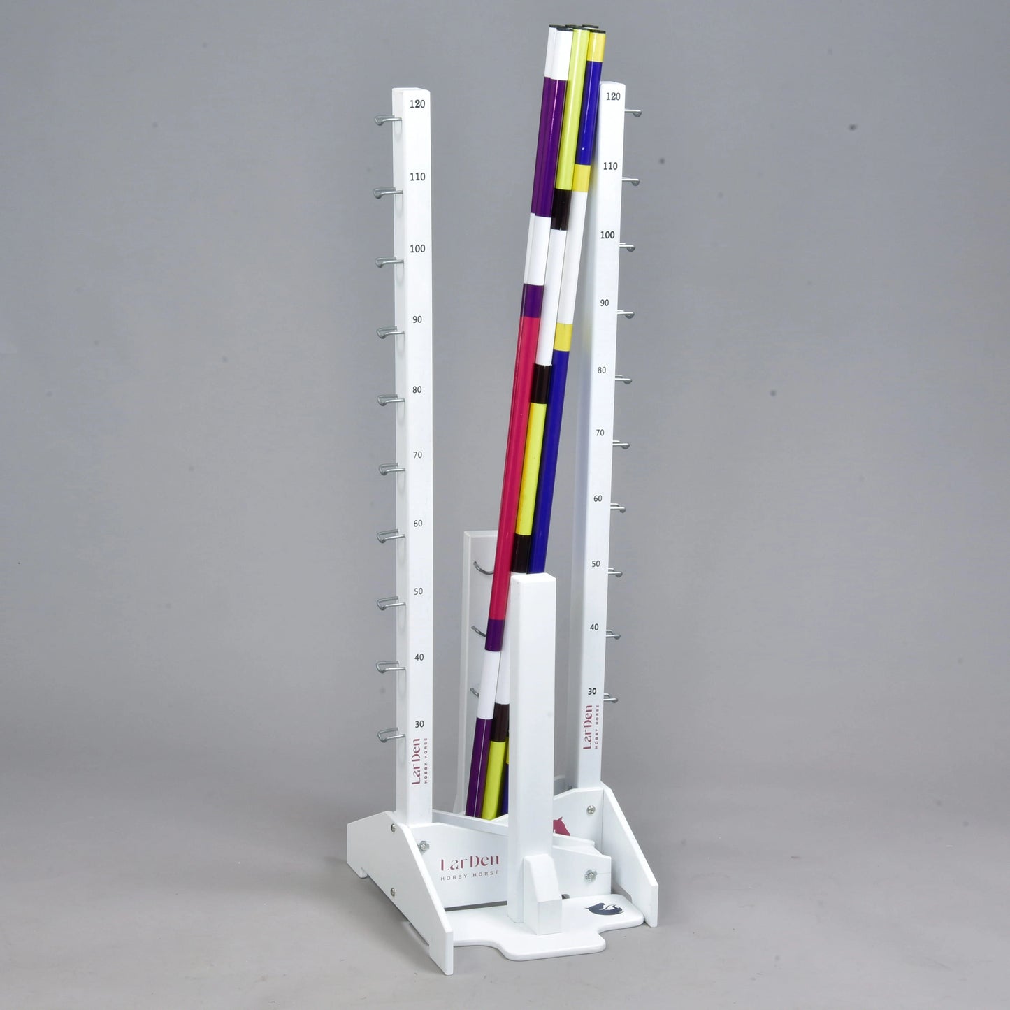 The Hobby Horse LarDen Jump set for Hobby Horsing features a vertical white stand with measurement markings and colorful striped poles in purple, yellow, white, and black—ideal for jumps or adjustable cavaletti. The background is plain gray.