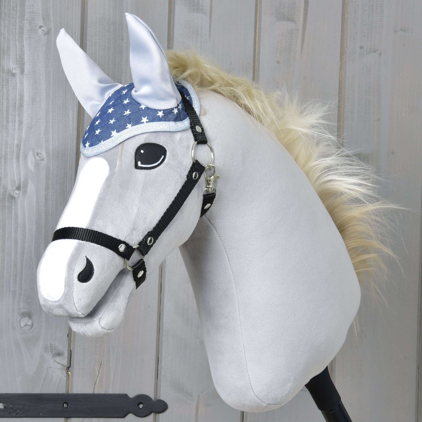 Nauszniki Star Hobby Horse LarDen to pluszowy Hobby Horse z białą głową, czarną uzdą, niebieskimi nausznikami w gwiazdy do ochrony przed owadami, jasną grzywą i oczami w stylu kreskówkowym, pokazany na drewnianym tle.