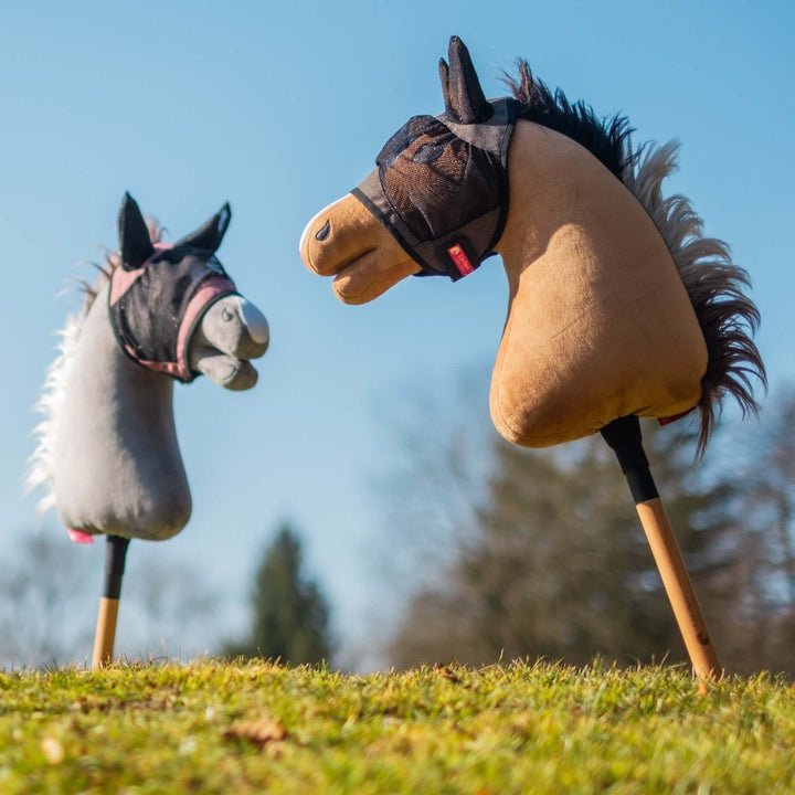 Dwie zabawki hobby horse z pluszowymi głowami, jedna szara, a druga brązowa, stoją pionowo na trawie na tle czystego błękitnego nieba i rozmytych drzew. Oba konie mają małe materiałowe uzdy, gotowe do pomysłowych przygód z hobby horsing.