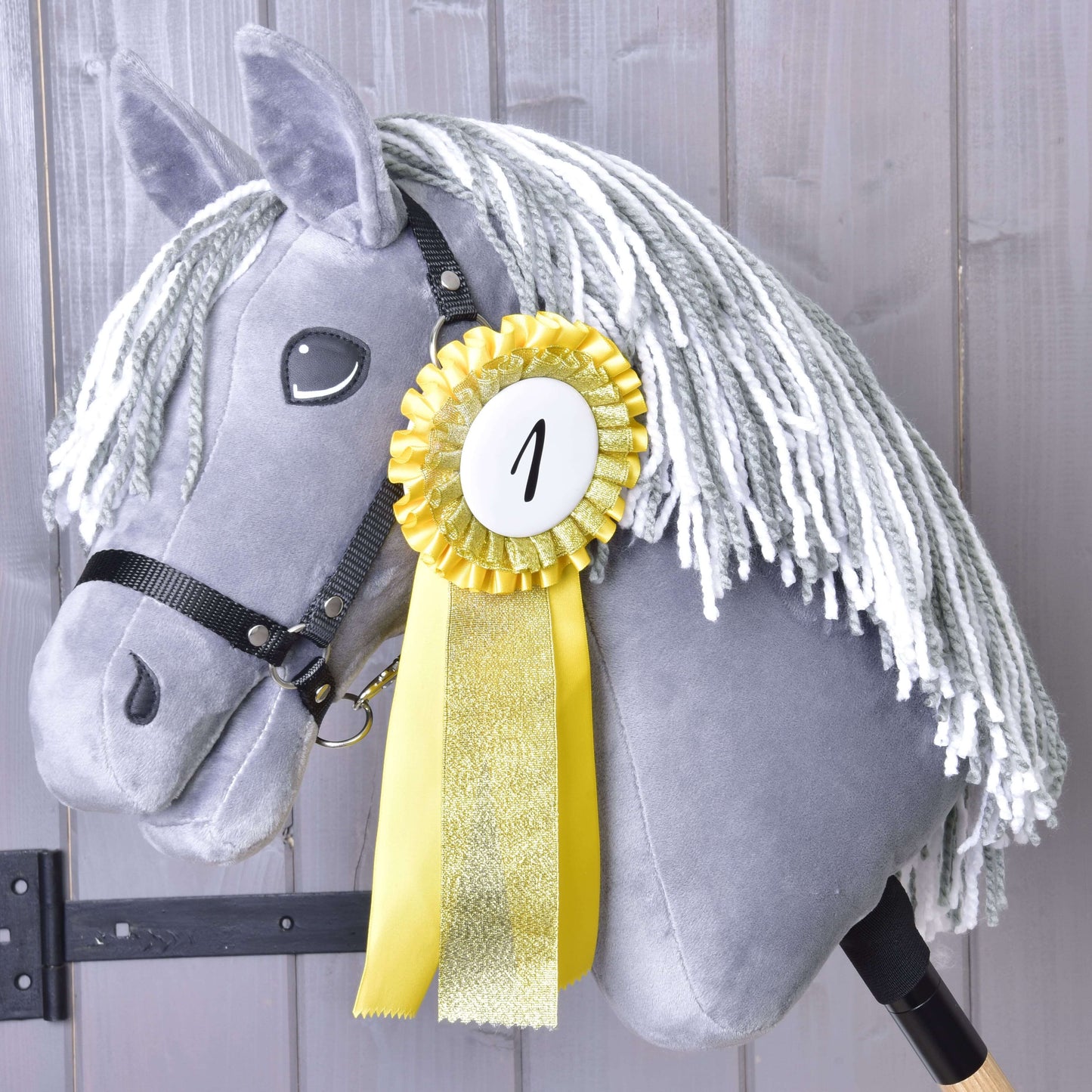 Zestaw Wstążek Hobby Horse NOWOŚĆ od Hobby Horse LarDen, z żółtą wstążką za pierwsze miejsce oznaczoną „1” na szarym Hobby Horse z białą grzywą z włóczki, pokazanym na tle drewnianej ściany.
