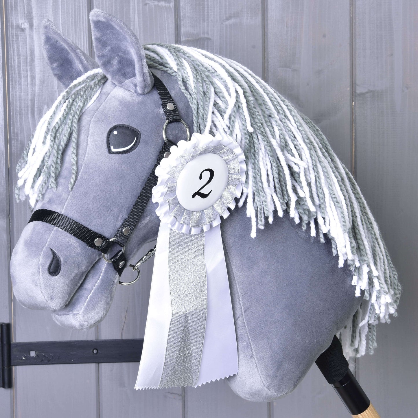 Szary Hobby Horse z białą grzywą z włóczki i czarną uzdą na drewnianym kiju prezentuje Zestaw Wstążek Hobby Horse NOWOŚĆ (drugie miejsce, srebrne wykończenie, numer 2) od Hobby Horse LarDen na tle szarych drewnianych desek.