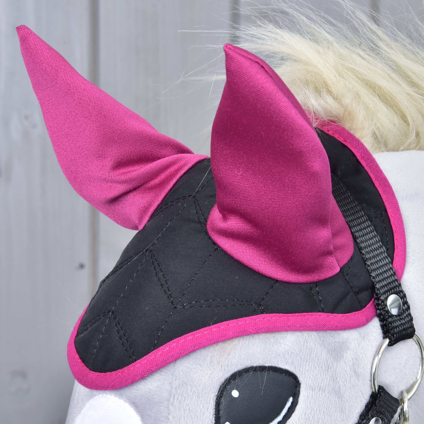 Close-up of a Hobby Horse LarDen Ear Bonnet Thermo Sky in black and magenta with two pointed magenta ears and a quilted pattern, providing insect protection, shown against a light wooden background.
