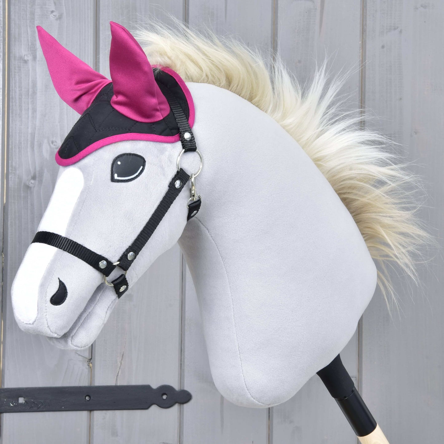 A gray Hobby Horse LarDen toy with a black bridle, white mane, and the bright pink Ear Bonnet Thermo Sky, designed for insect protection, posed in front of a wooden plank background.