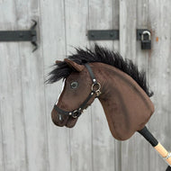 Hobby Horse Lucas z czarnym kantarem od Hobby Horse LarDen, pokazany na tle szarych drewnianych drzwi z czarnymi metalowymi zawiasami i zasuwką.