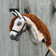 Hobby Horse Pompo z czarnym kantarem od Hobby Horse LarDen z szarym nakrapianym pyskiem, brązową grzywą z włóczki i czarną uzdą, inspirowany ogierem hanowerskim, pokazany na tle postarzanego drewnianego płotu.