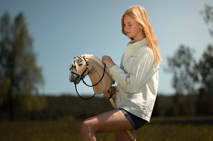 Młoda osoba z długimi blond włosami, w lekkiej kurtce i szortach, cieszy się hobby horsing, jadąc na hobby horse na słonecznej, trawiastej łące z drzewami w tle.