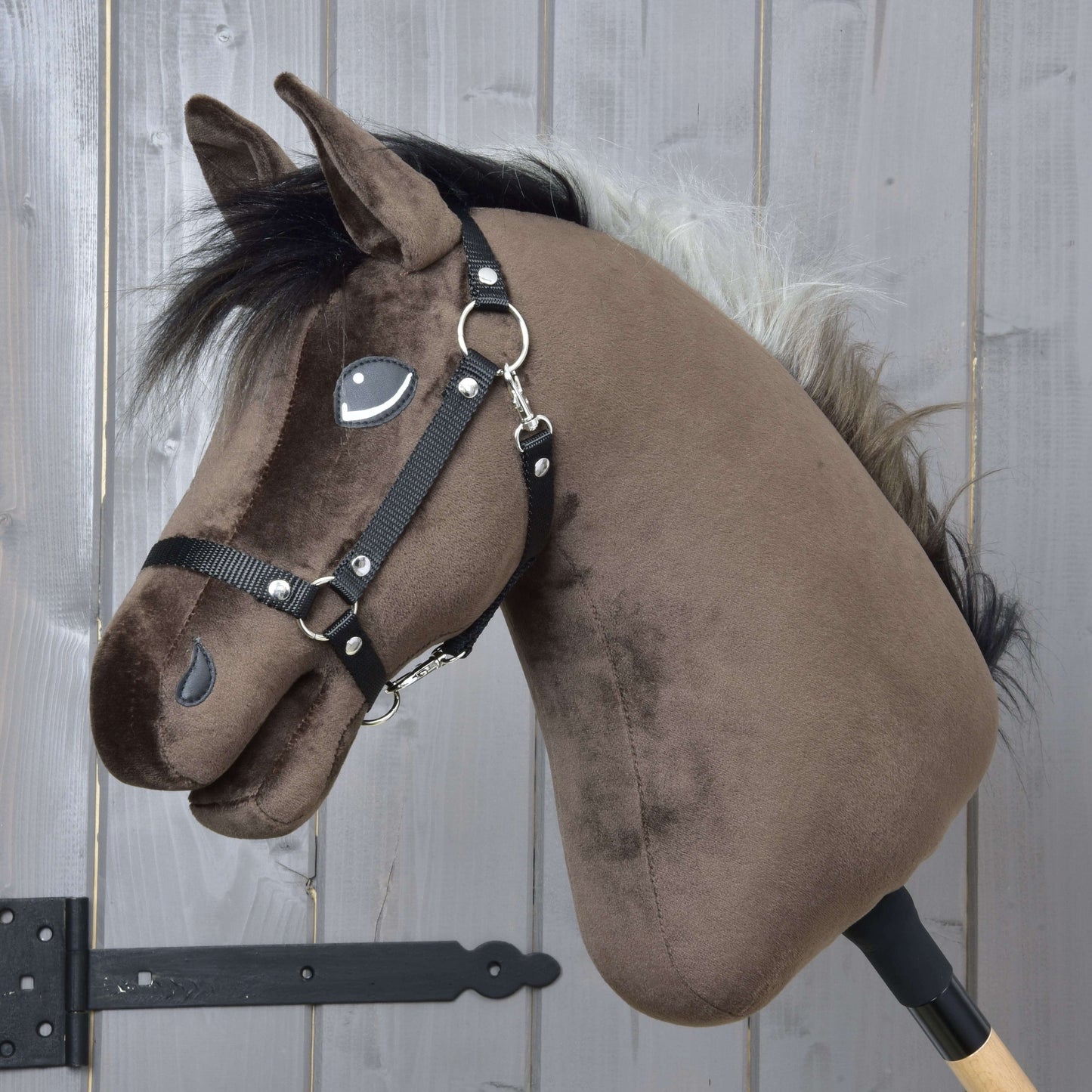 The Hobby Horse LarDen "Hobby Horse Alfredo with Black Halter" features a brown plush Thoroughbred-style head with black mane, white forelock, and black halter, mounted on a wooden stick against a gray wooden background.