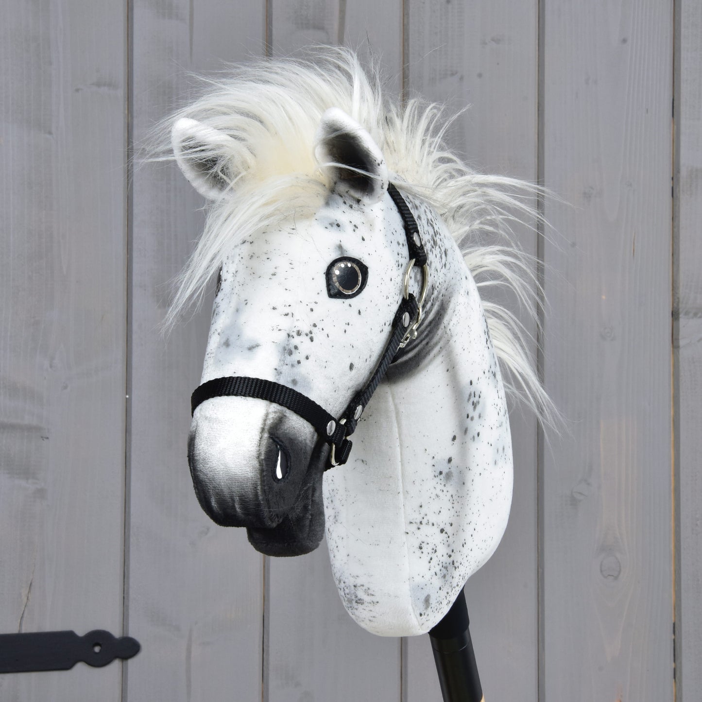 The Hobby Horse LarDen Aspen with Black Halter (size S), featuring white and black speckled markings and a fluffy mane reminiscent of an Appaloosa, is mounted on a stick and displayed against a wooden panel background.