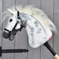 Hobby Horse Aspen z czarnym kantarem, rozmiar S od Hobby Horse LarDen, z białą pluszową głową ozdobioną czarnymi cętkami, puszystą grzywą i czarnym kantarem, osadzony na drewnianym kiju i pokazany na tle szarej drewnianej ściany.
